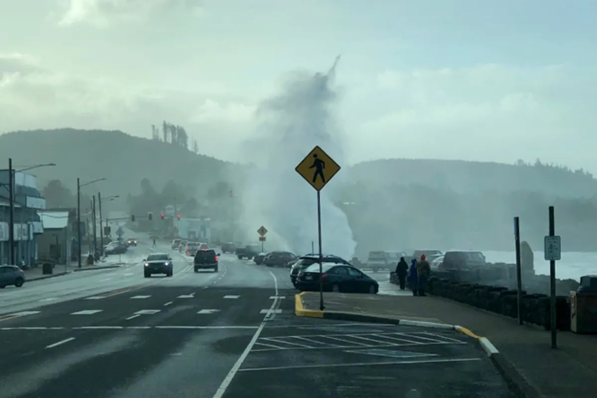 A tall plume of sea spray rising above the seawall beside the highway through Depoe Bay as people watch from the sidewalk