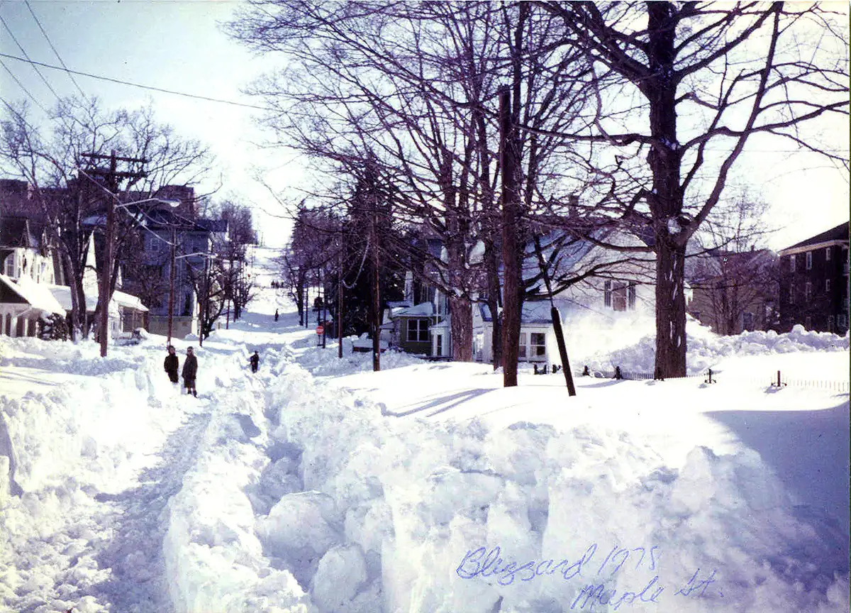 A residential street in Woonsocket walled in by high snowbanks, with a narrow shoveled lane and people walking between bare trees