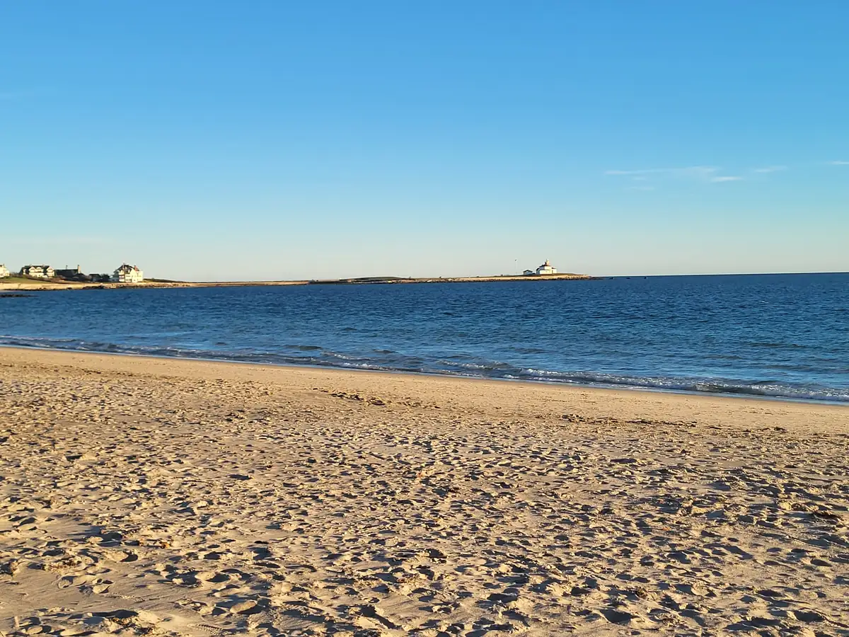 A wide sandy beach and calm blue water, with the white Watch Hill Lighthouse on a low point in the distance.
