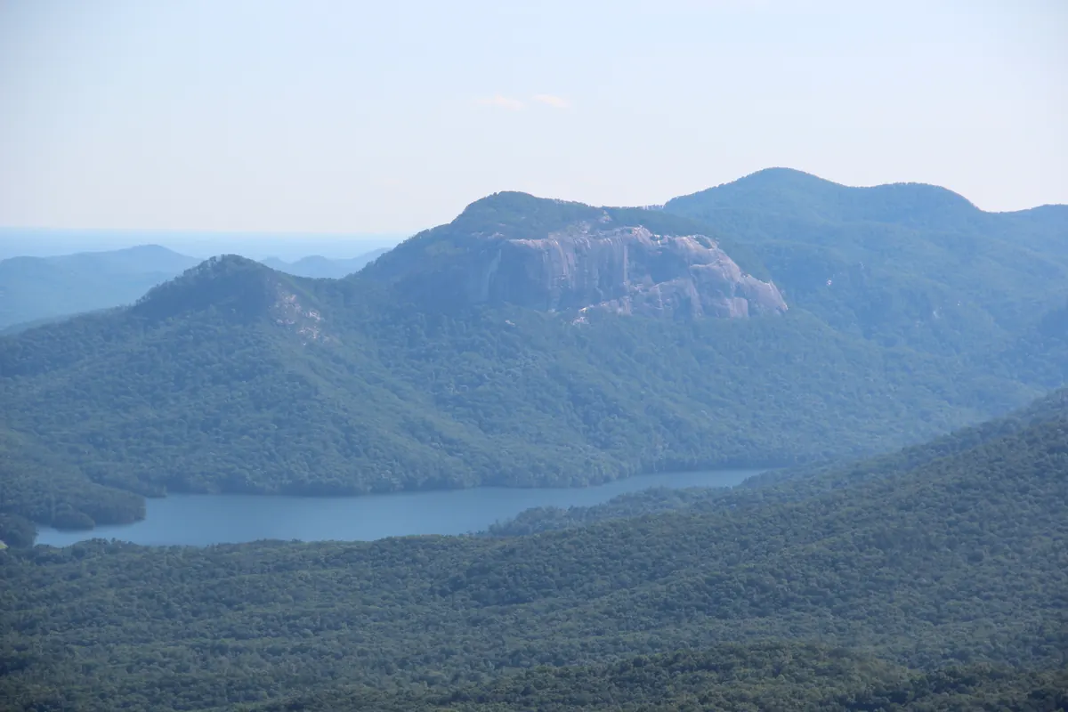 Forested mountains and the bare granite face of Table Rock seen from a high overlook on the Blue Ridge Escarpment, under a hazy sky.