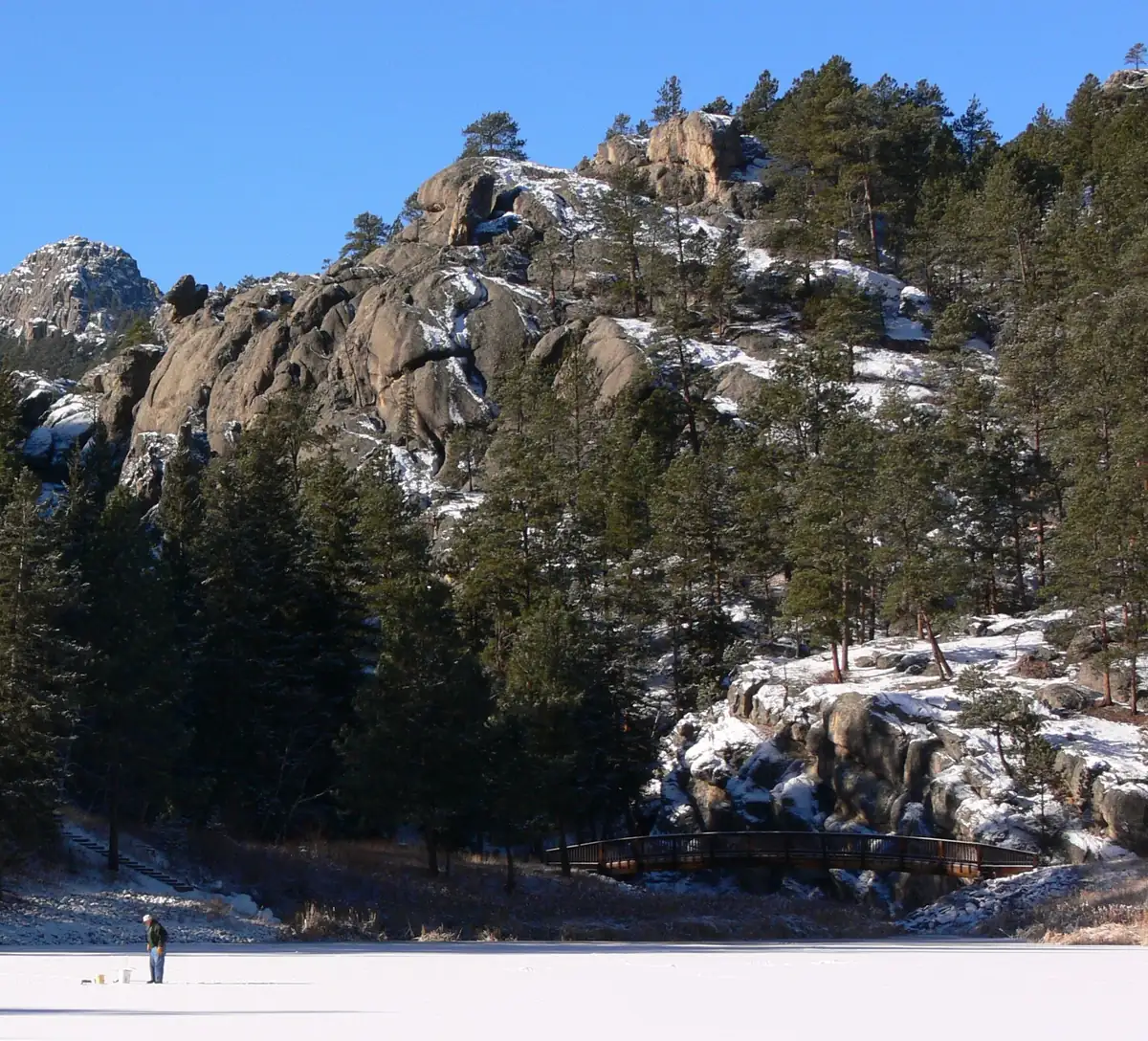 A lone angler stands on a snow-covered frozen lake below a snow-dusted granite ridge and ponderosa pines.
