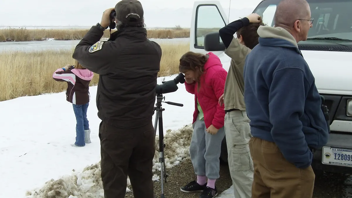A small group of adults and children with binoculars and a spotting scope looking out over a snowy marsh