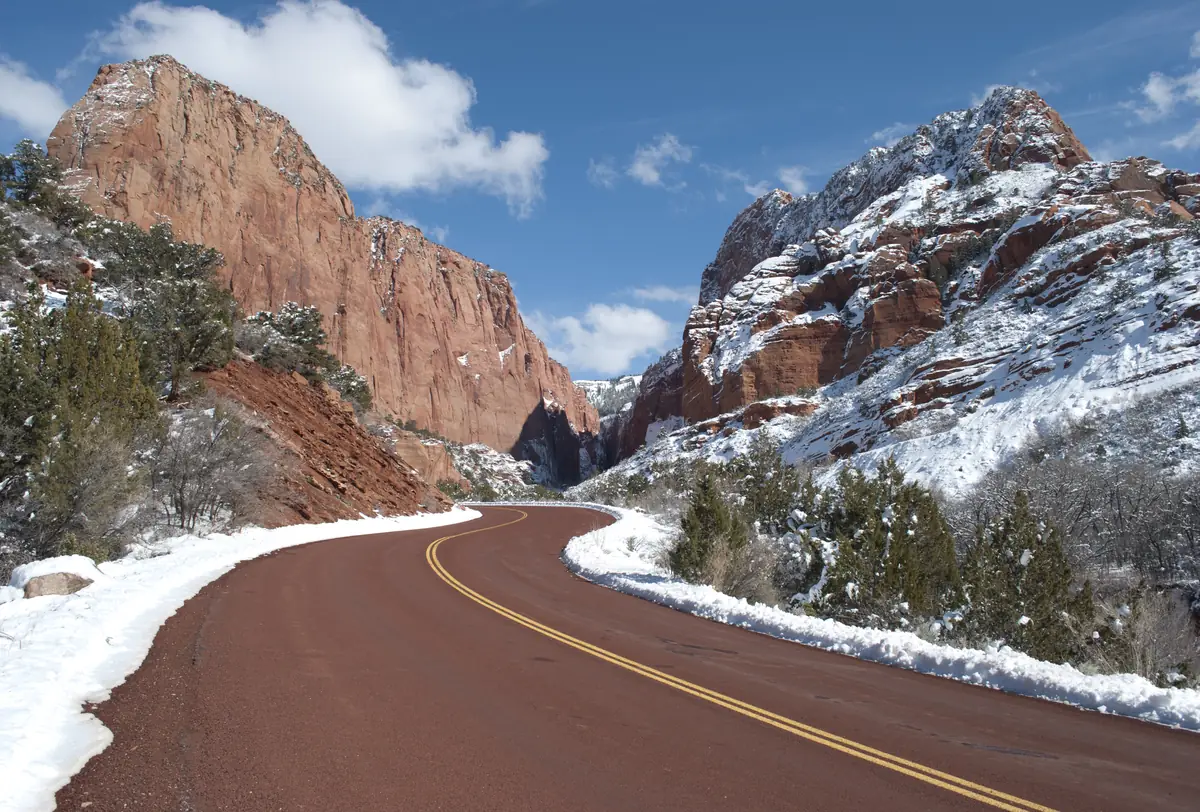 A red paved road curving between red sandstone cliffs and slopes dusted with fresh snow under a blue sky