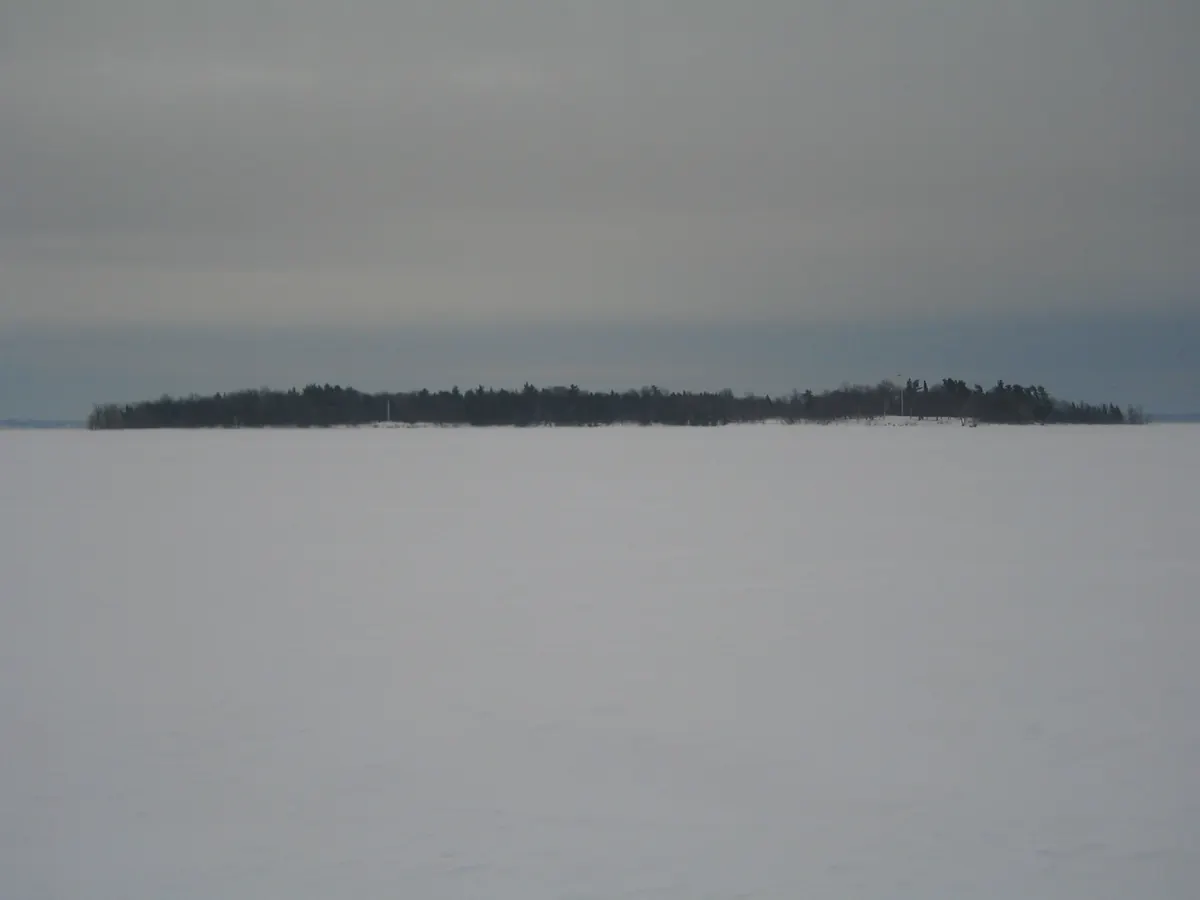 A flat white expanse of frozen lake under gray sky with a low wooded island on the horizon