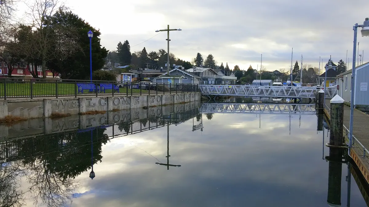 High water reaching almost to the top of a concrete seawall and walkway at a small marina under a cloudy sky.