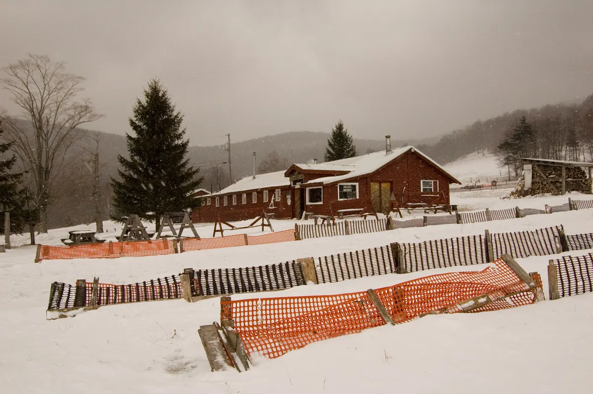 Rows of wooden and orange snow fences across a snowy field in front of a red lodge and evergreens, with snow falling over wooded hills.