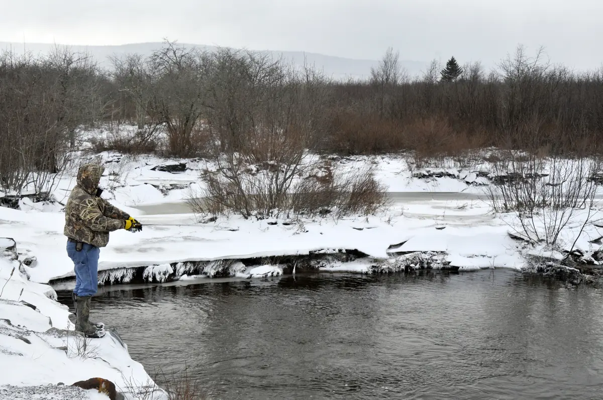 An angler in a camouflage jacket and jeans casts from a snowy bank into a dark, open stream lined with bare shrubs.