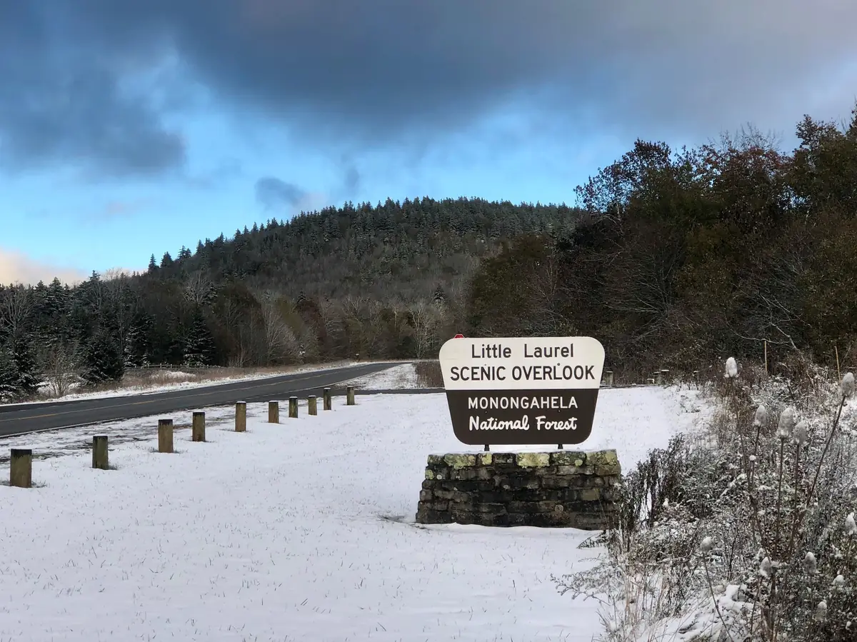 A light coat of snow on the grass and stone sign at Little Laurel Scenic Overlook, with spruce-covered hills and a gray sky behind.