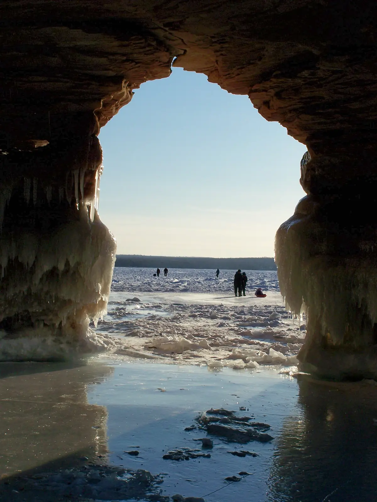View out of an icicle-lined sandstone sea cave toward visitors walking on the frozen surface of Lake Superior