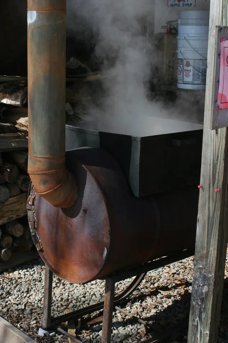 A steaming wood-fired evaporator pan on a rusty steel barrel stove with a stovepipe, stacked firewood behind it