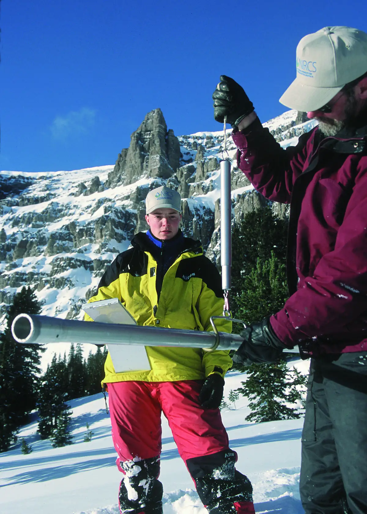 Two snow surveyors weighing a long metal snow sampling tube on a hanging scale below snowy cliffs