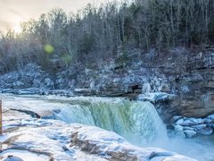 Cumberland Falls pours green-white water over a ledge between banks of snow and ice, with bare winter forest on the cliffs above.